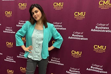 Aimen Khan wears gray pants, an aqua button down shirt and a teal jacket. She has her hands on her hips and is looking at the camera with her head tiled and a slight smile. In the background is a maroon CMU logo wall.
