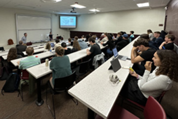 Students sit in a full classroom and watch a speaker at the front of the room as he delivers a presentation. There is a screen with the presentation slides in the corner.