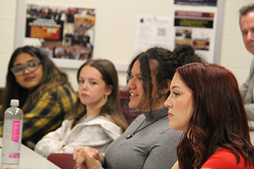 Four female students sit at a table during the inaugural AMA CMU chapter meeting. There is a water bottle on the desk and a bulletin board to their right side. ehind them, President MacKinnon listens intently.
