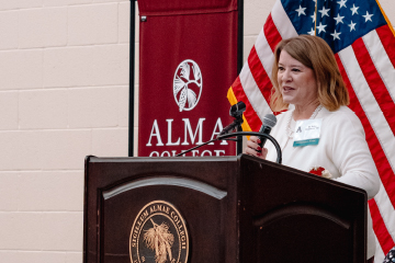 Amy McGinnis speaks into a handheld microphone behind an Alma College podium, with an American flag beside her.