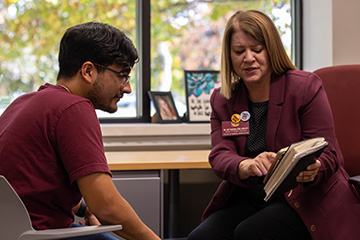 Amy McGinnis wears a maroon blazer and black shirt as she gestures to a notepad to show information to a student wearing a maroon t-shirt and glasses who sits opposite from her.