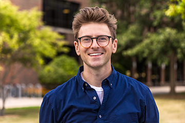 Anthony Niemiec wears a dark blue button down shirt that is open at the neck showing a white t-shirt. He has two-tone black and tortoiseshell glasses and his hair is combed up and back. He is smiling with Grawn Hall behind him.