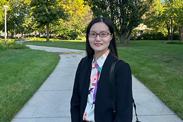 Ashley Yu stands on a sidewalk surrounded by green grass and trees. She has long black hair, brown oval frame glasses and red lipstick. She is wearing a black jacket with a vibrant shirt with splashes of pink, orange, teal and purple.