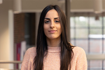 Tamara Merseli wear a peach ribbed mock turtleneck sweater and looks at the camera, smiling slightly. She has long dark brown hair. She's standing in Grawn Hall, but the background is blurry and hard to identify.