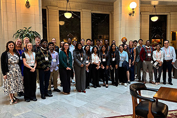 More than thirty students and faculty members stand in an upscale atrium with marble floors and stone columns. The are dressed in business professional clothing and wear orange lanyards with nametags.