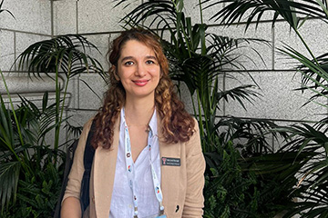 Bahar Javadi Khasraghi stands in stone hallway in front of palms and ferns. She has a tan jacket with a name tag, a conference badge, and a purse over her shoulder. She's smiling at the camera.
