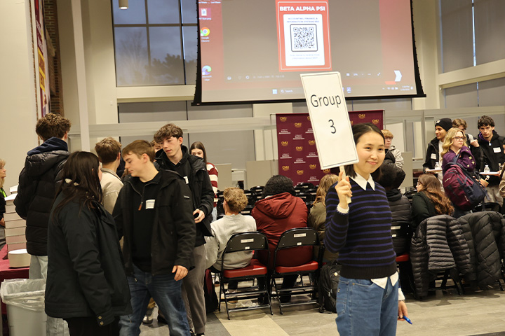 Students sit in chairs in the Grawn atrium while others form a queue to grab pizza. In the foreground, a student in a blue sweater and jeans holds a sign that reads 