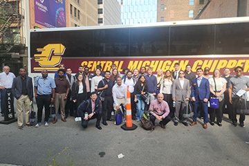 A large group Beta Alpha Psi members, accounting and finance students and faculty members wear business professional clothing and stand in front of a CMU branded bus. The bus is parked on a Detroit city street, there is an orange and white traffic cone.