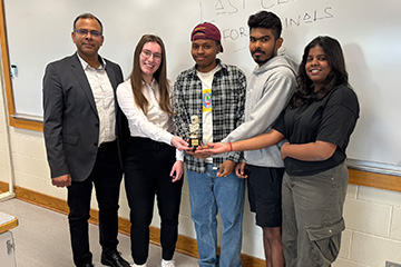Associate Dean Sanjay Kumar stands with the I-Ride student team in a classroom as they hold an award together. The group is positioned in front of a whiteboard, representing their project work in BIS 636.