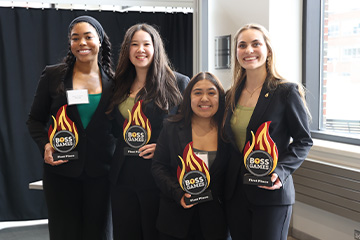 Four students hold first-place Boss Games awards and smile at the camera during a College of Business competition event.