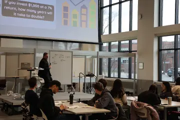 Rudy Bustamante sits on a Grawn Atrium stage in front of a white board with equations while students sitting at tables work through the financial resource exercises.