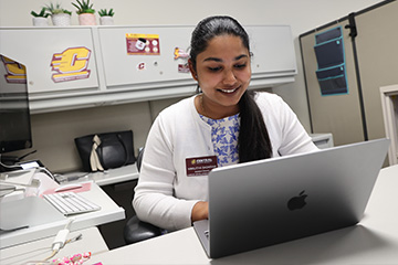 Nimilitha Narayani Sadaram has long black hair that is pulled into a pony tail. She sits at a desk in her office facing the camera and looks at her silver laptop smiling.