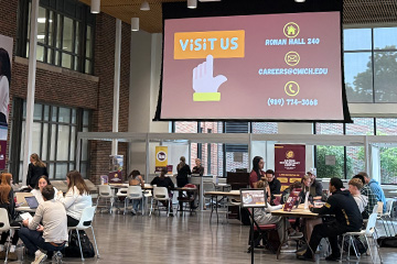 Students in business clothing sit at tables in the Grawn Atrium. The overhead screen has a slide that has a hand icon pointing toward the words "Visit us" and the information for the Career Development Center.
