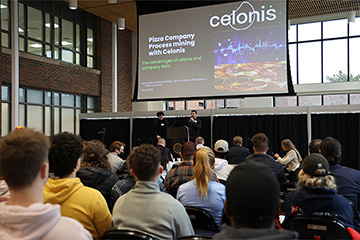 Students sit in the Grawn Hall atrium facing a lifted stage with two speakers presenting at a Celonis process mining competition. A display screen is behind and above them with a slide that reads "Pizza Company Process mining with Celonis."