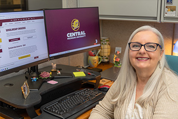 Cindy Howards sits at her desk with the Scholarship Universe web page displayed on her computer monitor. She has long white and gray hair, black glasses and silver earrings. She's wearing a tan cardigan over a white tank top.