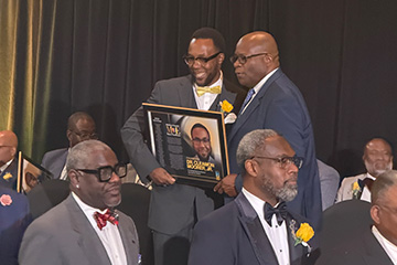 Management faculty member Cleamon Moorer Jr. accepts a 2026 Michigan Chronicle Men of Excellence award while holding a framed recognition plaque during the awards ceremony.