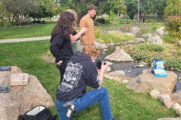 Four students stand around a game box for Hydrologic Cycle which has been posed on a rock in the outdoor gardens on the CMU campus. The students have cameras and phones and are taking photos for the game's publisher.