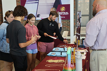 Prospective students and parents face an information table with giveaways like blue drawstring bags and colorful necklaces. A faculty member stands behind the table and speaks with them. In the background is another information table.