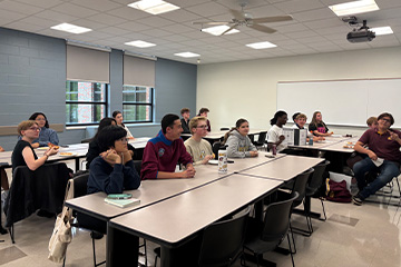Students sit at classroom tables with pizza and drinks, looking toward someone outside the frame.
