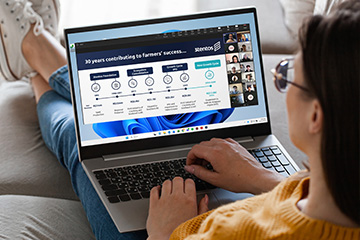 A student sits on a couch with a laptop on her lap. On the screen is a PPT slide with content from Cristiano Machado Costa's presentation. Students appear in the participant gallery on the screen as well.