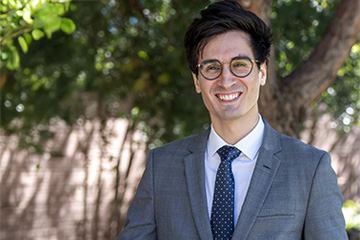 Daniel Belchior stands in front of a tree. He is smiling and waring a gray suit jacket over a white shirt and blue tie with white polka dots. He has black framed glasses.
