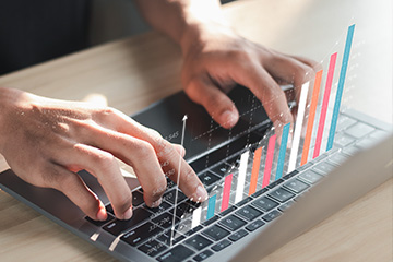 Two hands type on a laptop keyboard that is sitting on a light natural wooden tabletop. A virtual reality style bar graph hovers over the keyboard showing bars in white, blue, red and orange on a white graph.