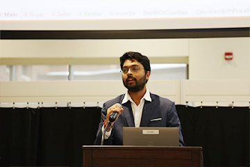 Shiva Karthik Rallabandi stands on a stage in the Grawn Atrium. He wears a blue gray suit with a white shirt and white pocket square. He holds a microphone and stands at a podium with a laptop.