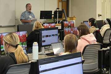 Charles Parker stands in front of a classroom lab with students sitting at desktop computers. He is wearing a blue and gray patterned shirt and holding a packet of papers at the front of the room while students listen to him speak.