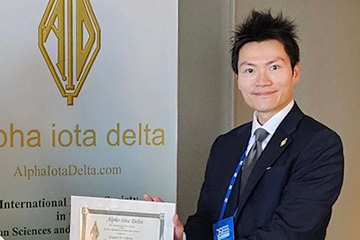 Edward Chou poses in front of an Alpha Iota Delta banner at the annual meeting. He has black spiky hair and is wearing a suit jacket with a white button-down shirt and a gray tie. He is holding an Alpha Iota Delta certificate.