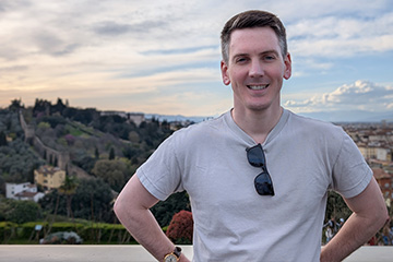 Adam Effinger stands outside in front of a European vista of hills and a small town. He wears a gray t-shirt with sunglasses hanging from the neckline.