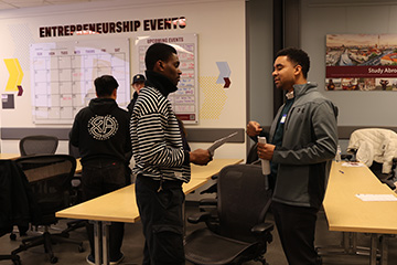 Two students in jackets stand in the Grawn 150G classroom. There are students talking, tables, and the ENT calendar of events behind them.