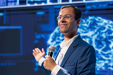 An entrepreneur gives a presentation holding a microphone. He is wearing a navy blazer and white shirt. He has glasses. Behind him is a digital display with neural pathways.