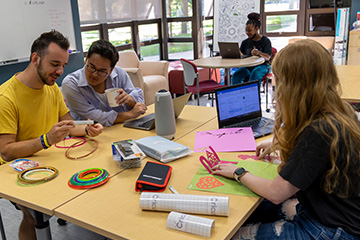 Students sit at tables in the IBIE Idea Den in Grawn Hall. In the foreground, three students look closely at 3D printed components and paper designs. On the table are prototyping supplies and a laptop. A student works on a laptop in the background.