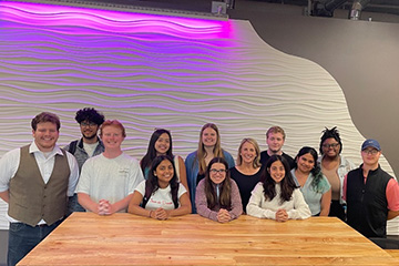 Twelve students and their advisor Julie Messing stand at a natural wood table. The students are of mixed genders and races. Behind them is a white textured wall with ripples and a neon purple light.