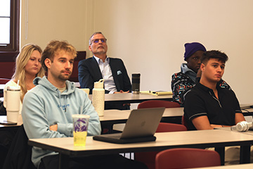 Jeff Hoyle sits in the third row of a classroom. He is wearing a dark suit jacket with a light blue pocket square. Five students fill the seats in front of him. They are all looking forward intently at something happening in the front of the class.