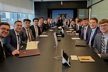 A group of 20 finance students sit in business professional clothing at a long conference table in a boardroom. They have notebooks and pens in front of them and all face towards the camera smiling.
