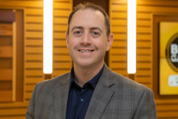 Fred Lawrence wears a gray plaid suit jacket over a black button down shirt. He has short brown hair and is standing in front of a wood slat wall in Grawn Hall.