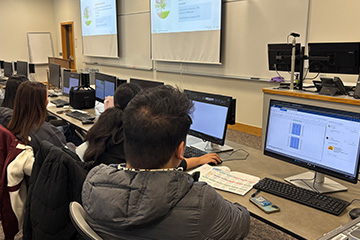 Team members in the global ERPsim West competition site at computer terminals in a computer lab in Grawn Hall. On the monitors are various spreadsheets and documents, as well as the SAP interface. They intently converse and strategize.