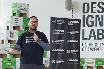 Gustav Verhulsdonck wears a black shirt with white letters that reads faculty innovation fellows over a long-sleeve gray shirt and gray pants. He has short brown hair and scruffy facial hair. He stands in front of a tower of green and white boxes.