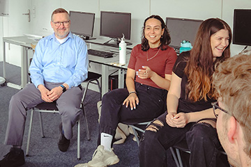 James Melton sits with a small group of students at Hochschule Karlsruhe in Germany. He is wearing gray pants, a light blue button down shirt, and an Apple watch. There are computers on desks in the classroom. The students are laughing and gesturing.