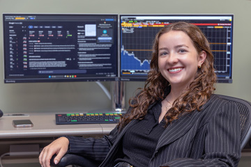 Holly Ireton sits in front of two computer monitors with market and financial data. She's wearing a gold necklace, a black and gray pinstriped blazer over a black shirt. She has long brown curly hair, blue eyes and a big smile.