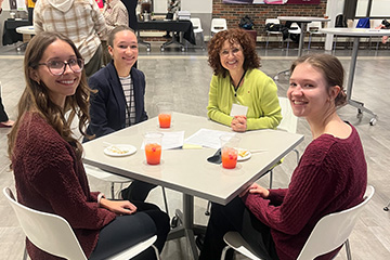 Three students in business clothing sit with a faculty member at a table in Grawn Atrium. They are all smiling at the camera. On the table are mocktails and plates.