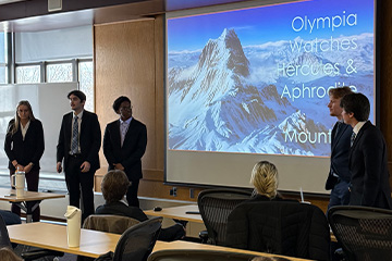 Five students stand at the front of a conference room wearing business professional clothing. They are presenting their iCore project. Behind them in a display screen with a PPT slide showing their smartwatch brand.