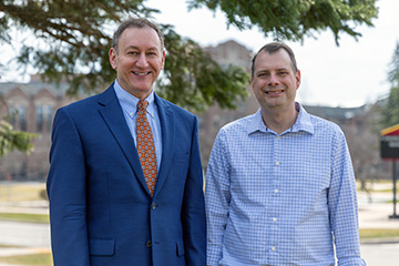 Adam Epstein (left) and Jason Taylor stand outdoors on Central Michigan University’s campus, smiling at the camera with campus buildings and trees in the background.