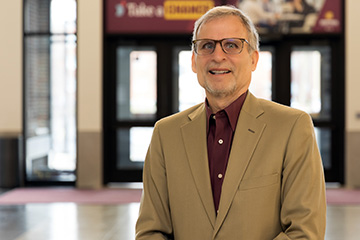 Jeffrey Hoyle, marketing and professional sales faculty member at Central Michigan University, stands smiling in the Grawn Hall Atrium wearing a tan blazer and maroon shirt.
