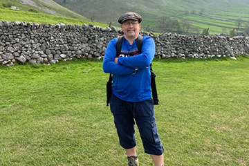 Jerry DiMaria wears a blue long-sleeve tshirt, blue cargo pants, hiking boots, backpack and cap. He's standing in a grassy field and there is a stone wall behind him with hills rising in the distance.