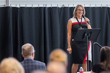 Julie Messing stands at a podium speaking into a microphone during a College of Business Administration event at Central Michigan University. Audience members are visible in the foreground as she addresses the crowd.