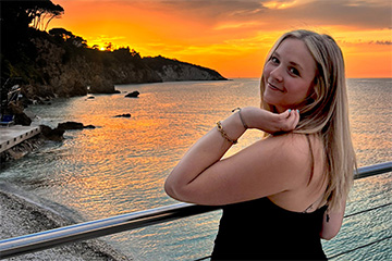 Kaitlyn Klumpp smiles beside a railing overlooking a rocky coastline and the sea at sunset.