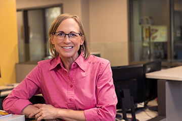 Karen Arthur has chin length blonde hair and is wearing black framed glasses and a bright pink button down shirt. She's standing at the front desk of the Business Student Services office.