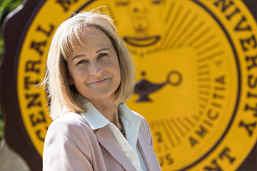Kathy Litwiller smiles in front of a large yellow and maroon Central Michigan University seal, wearing a light pink blazer over a white blouse.